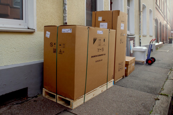 Verpackte Wärmepumpe und Anlagenteile auf Paletten vor einem Altbau, daneben eine Sackkarre.