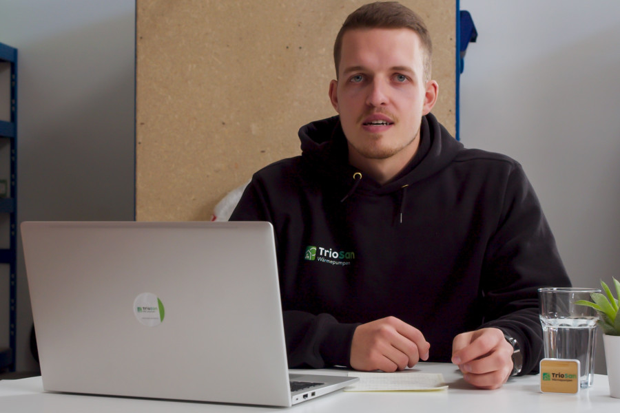 Torben von TrioSan im Firmen-Hoodie am Tisch mit Laptop, im Hintergrund das Lager.