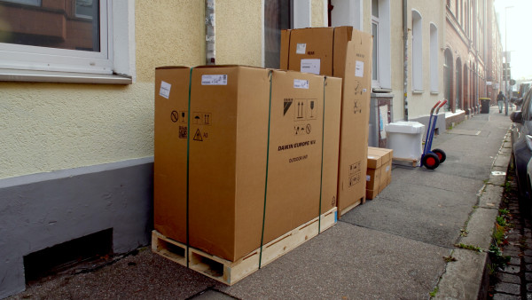 Verpackte Wärmepumpe und Anlagenteile auf Paletten vor einem Altbau, daneben eine Sackkarre.