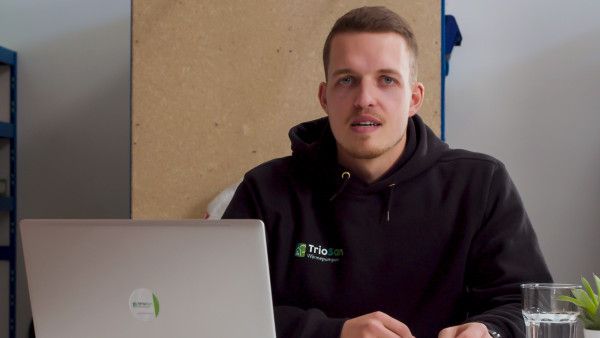 Torben von TrioSan im Firmen-Hoodie am Tisch mit Laptop, im Hintergrund das Lager.